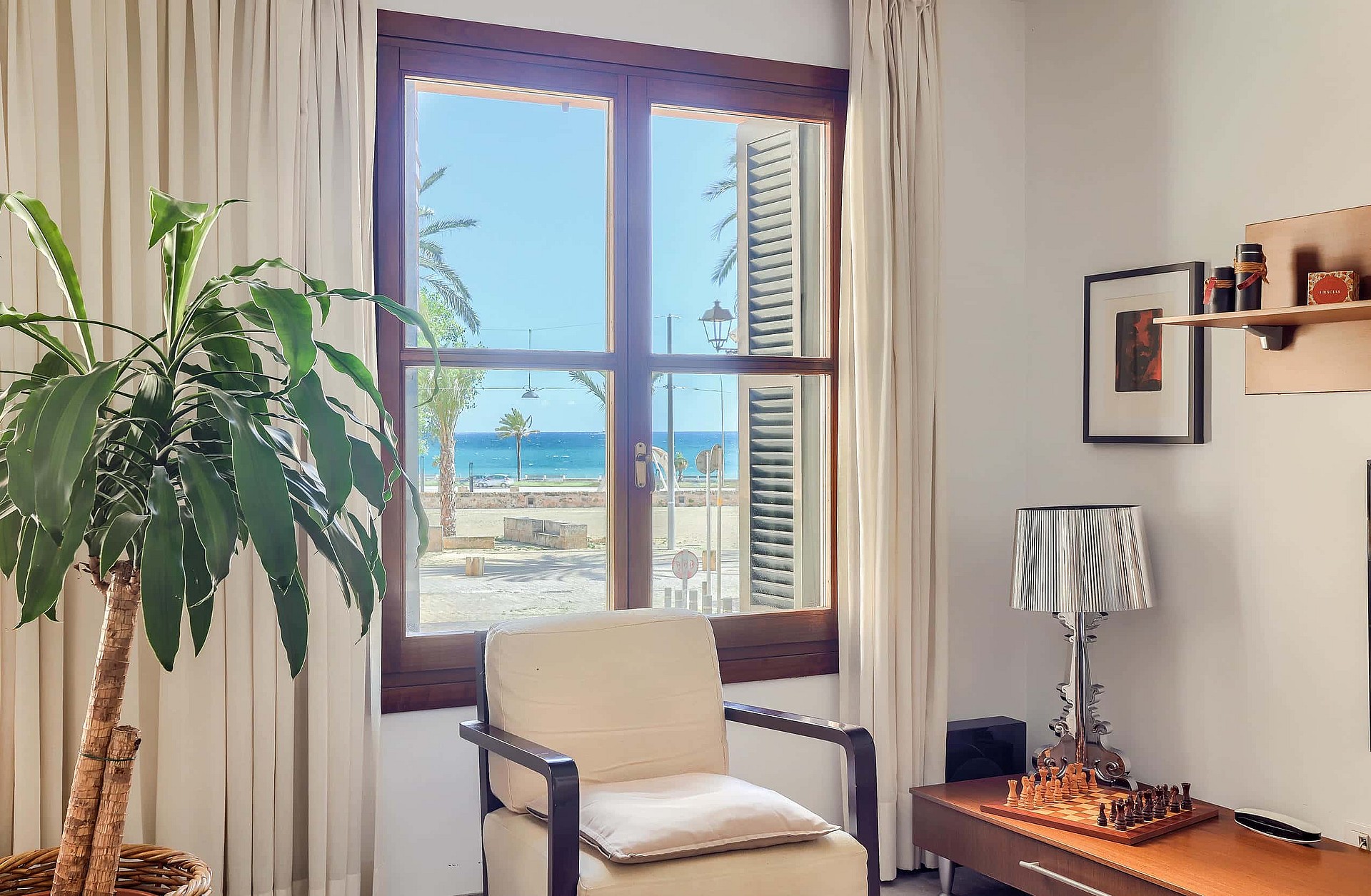 A bright living space with a wooden window frame looking out to the sea and palm trees. An armchair is placed near the window next to a houseplant and a side table with a lamp.