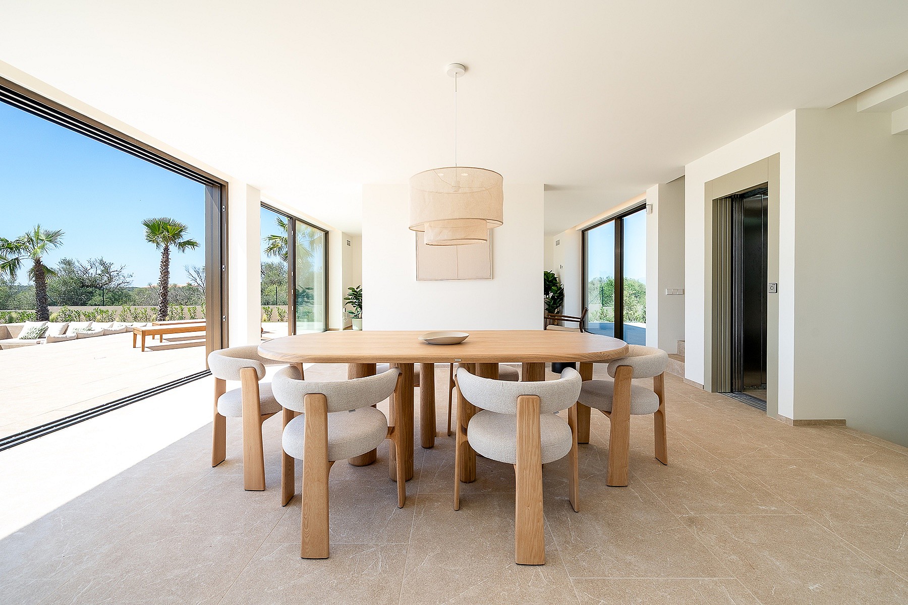 Dining room with light oak table and textured cream chairs, floor-to-ceiling glass walls opening to a sunlit terrace.