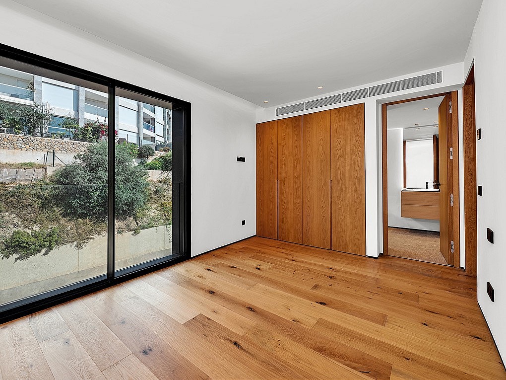 A minimalist bedroom featuring light oak flooring, large integrated oak wardrobes, and a sliding glass door looking out to greenery.