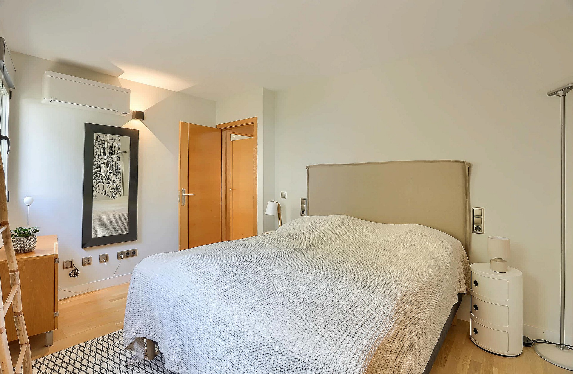 A bedroom featuring a double bed with a neutral headboard, textured white bedding, wooden door and cabinet, and wall-mounted air conditioning.
