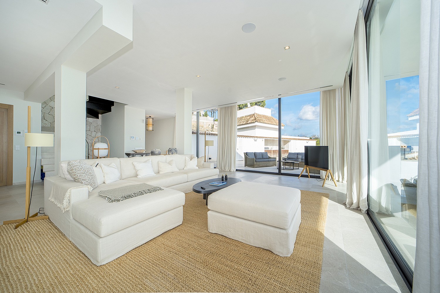 Bright living room with white L-shaped sofa, matching ottoman, woven area rug, and floor-to-ceiling glass sliding doors.