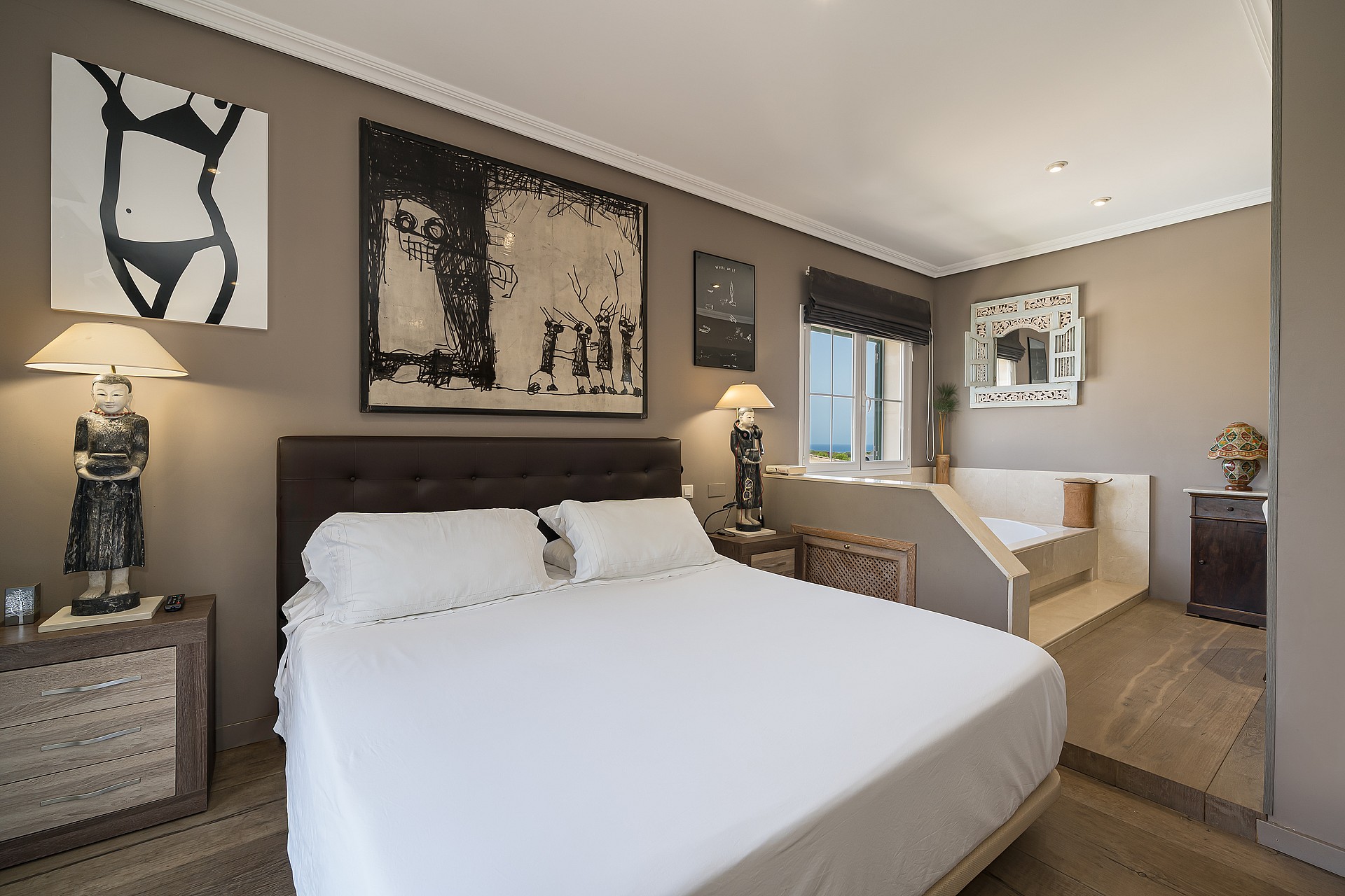 Bedroom with queen-sized bed, taupe walls, and an elevated bathroom area with a white soaking tub visible behind a low wall.
