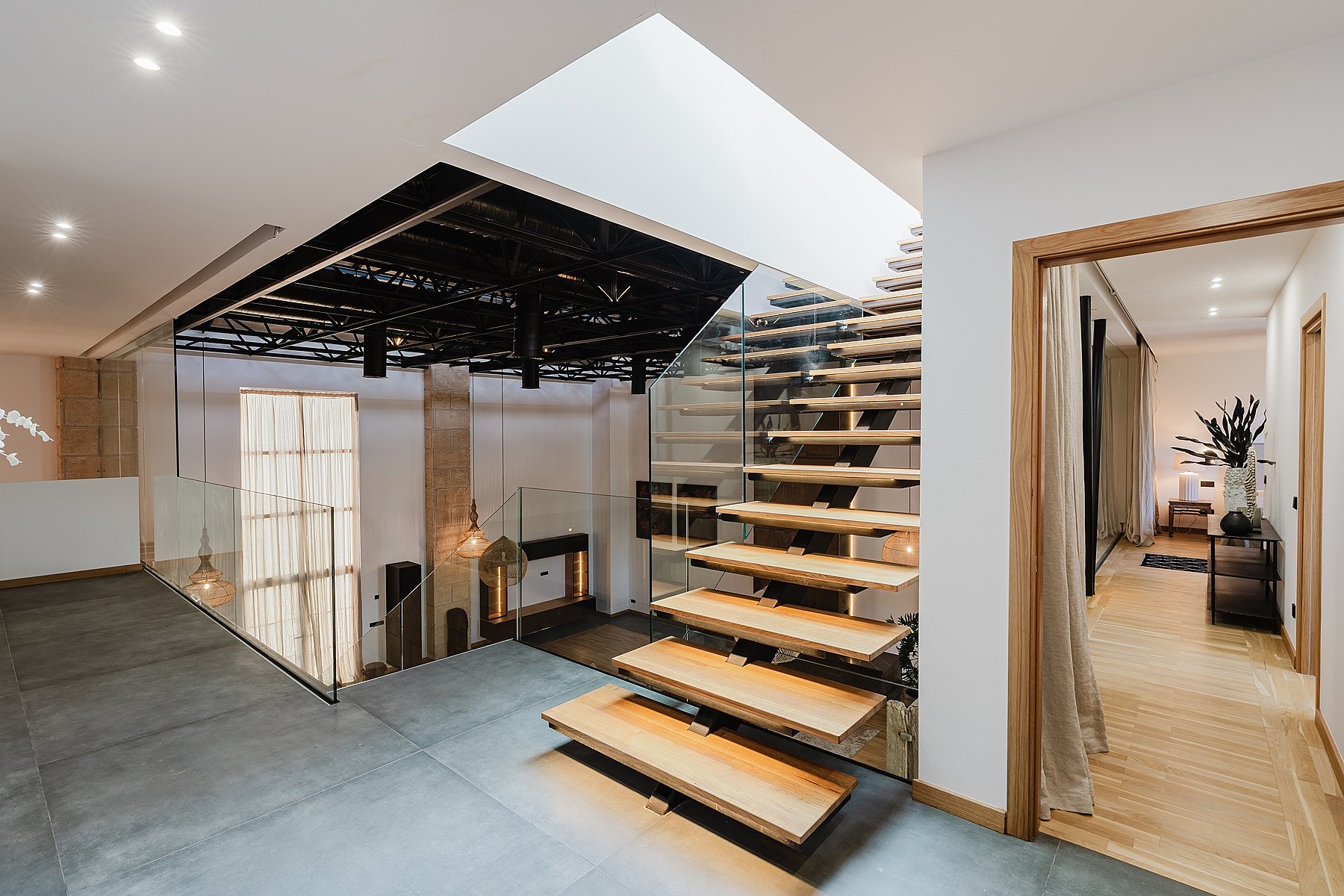 A modern floating oak staircase with glass side panels, illuminated by ceiling spotlights and a skylight.