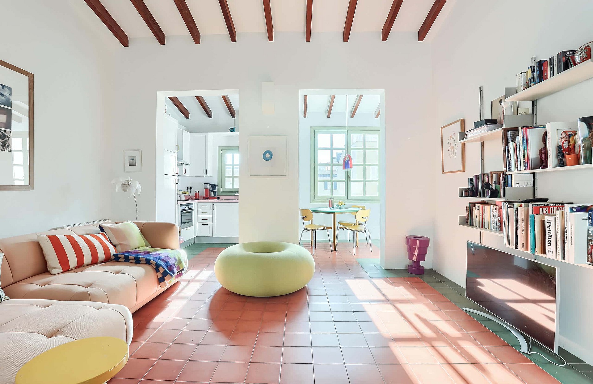 A bright living area featuring terracotta floor tiles, exposed wooden ceiling beams, a sofa with a striped cushion, and a round lime green ottoman, looking through to a dining area.