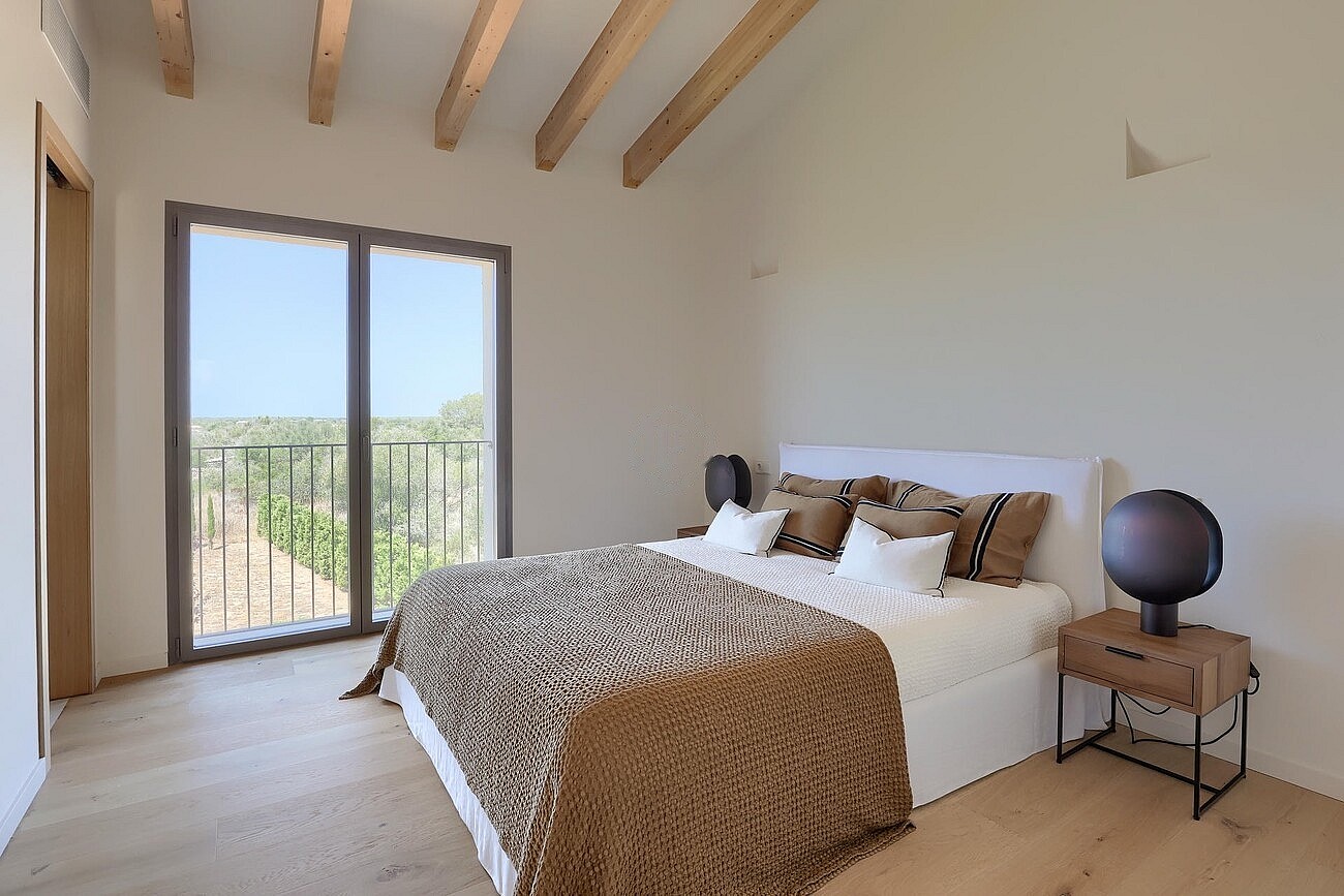 A bright bedroom featuring a queen bed with animal print bedding, light wood floors, and exposed ceiling beams with a balcony.