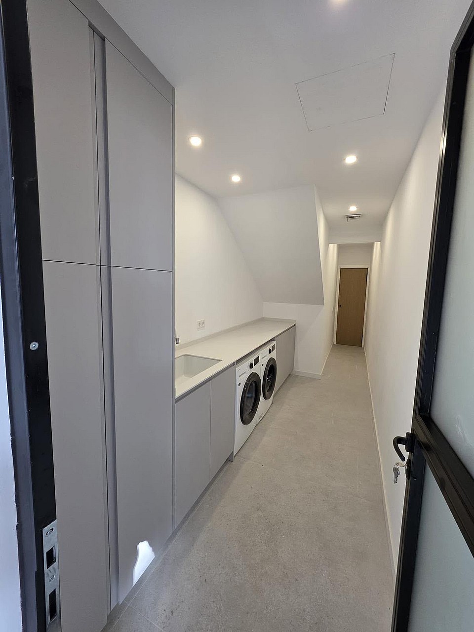 A narrow, minimalist laundry room with white cabinetry, integrated appliances, and light grey tile flooring.