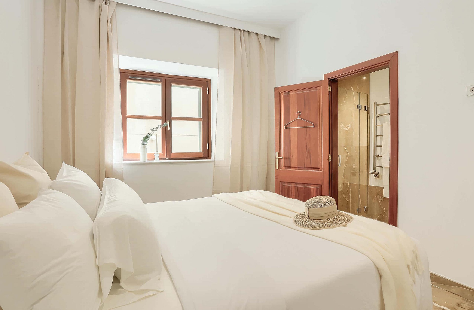 A bedroom interior with a white bed, beige curtains, a wooden window frame, and an open wooden door leading to a bathroom.