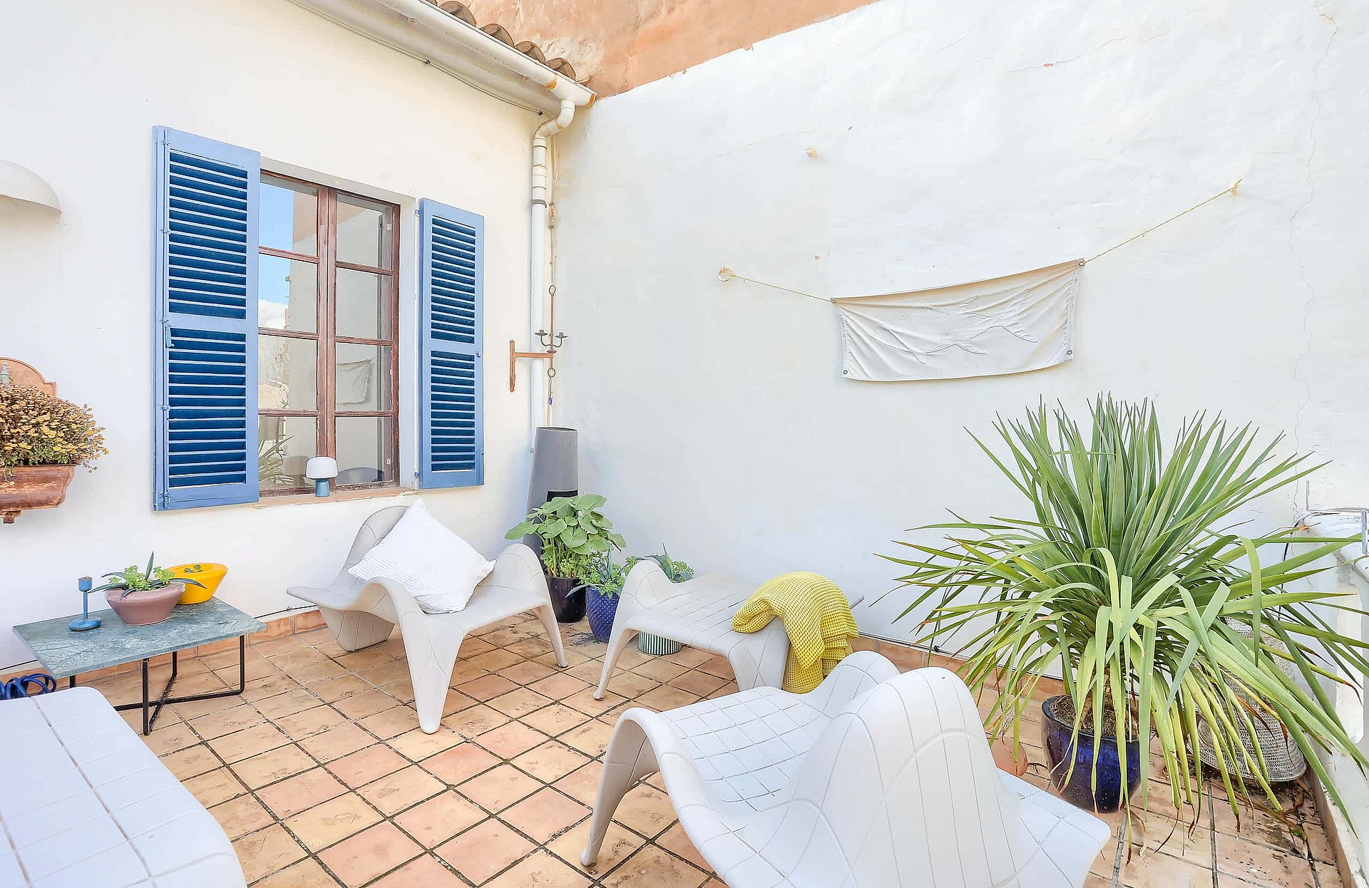 A small outdoor terrace with terracotta tiles, white plastered walls, blue wooden shutters, and modern white lounge chairs.