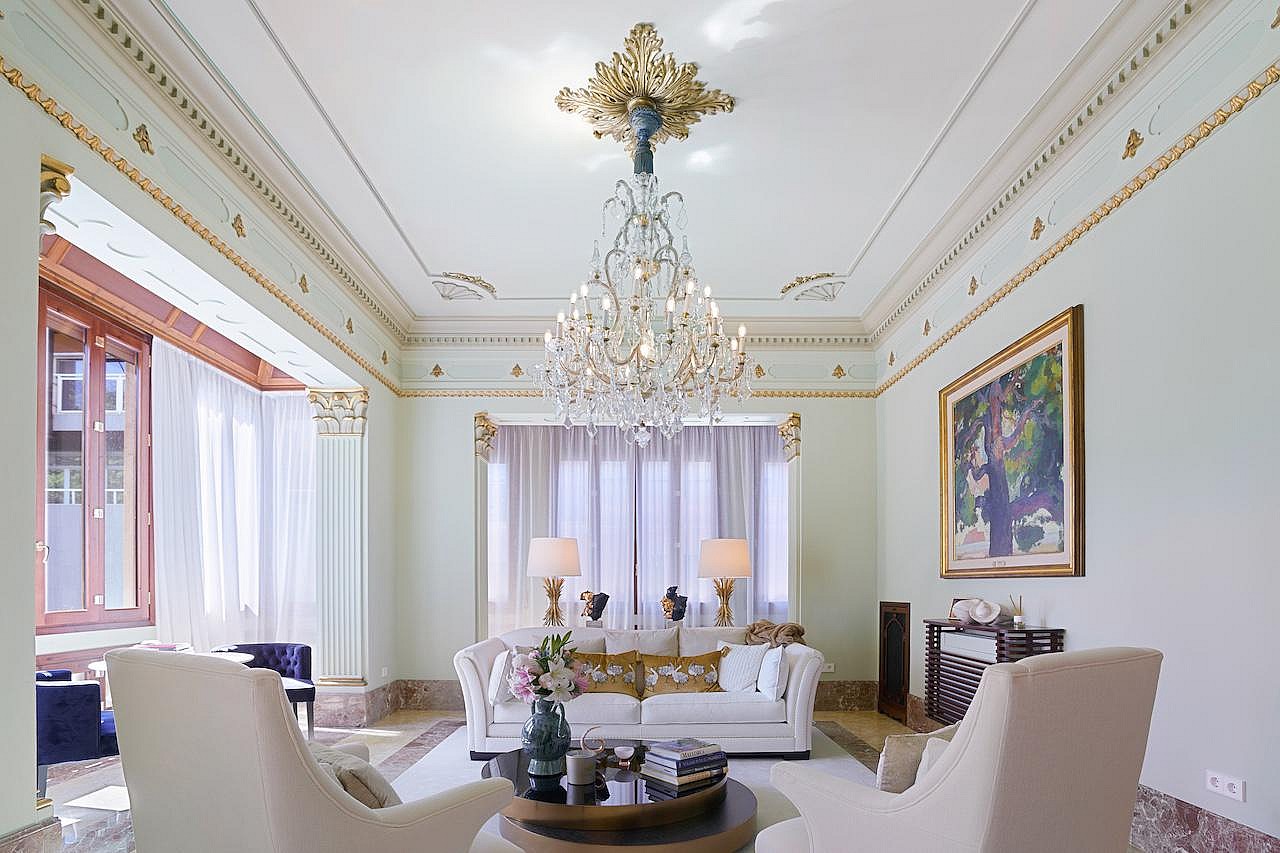 A formal living room with high ceilings, decorative gilded molding, and a crystal chandelier above a white sofa.
