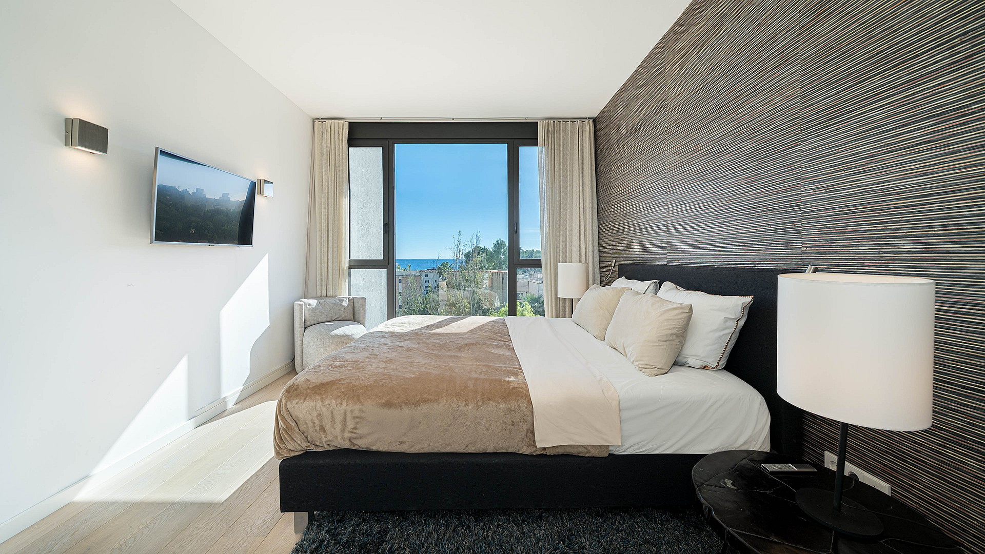 Modern master bedroom with king-sized bed, textured dark feature wall, and full-height windows overlooking a coastal scene.