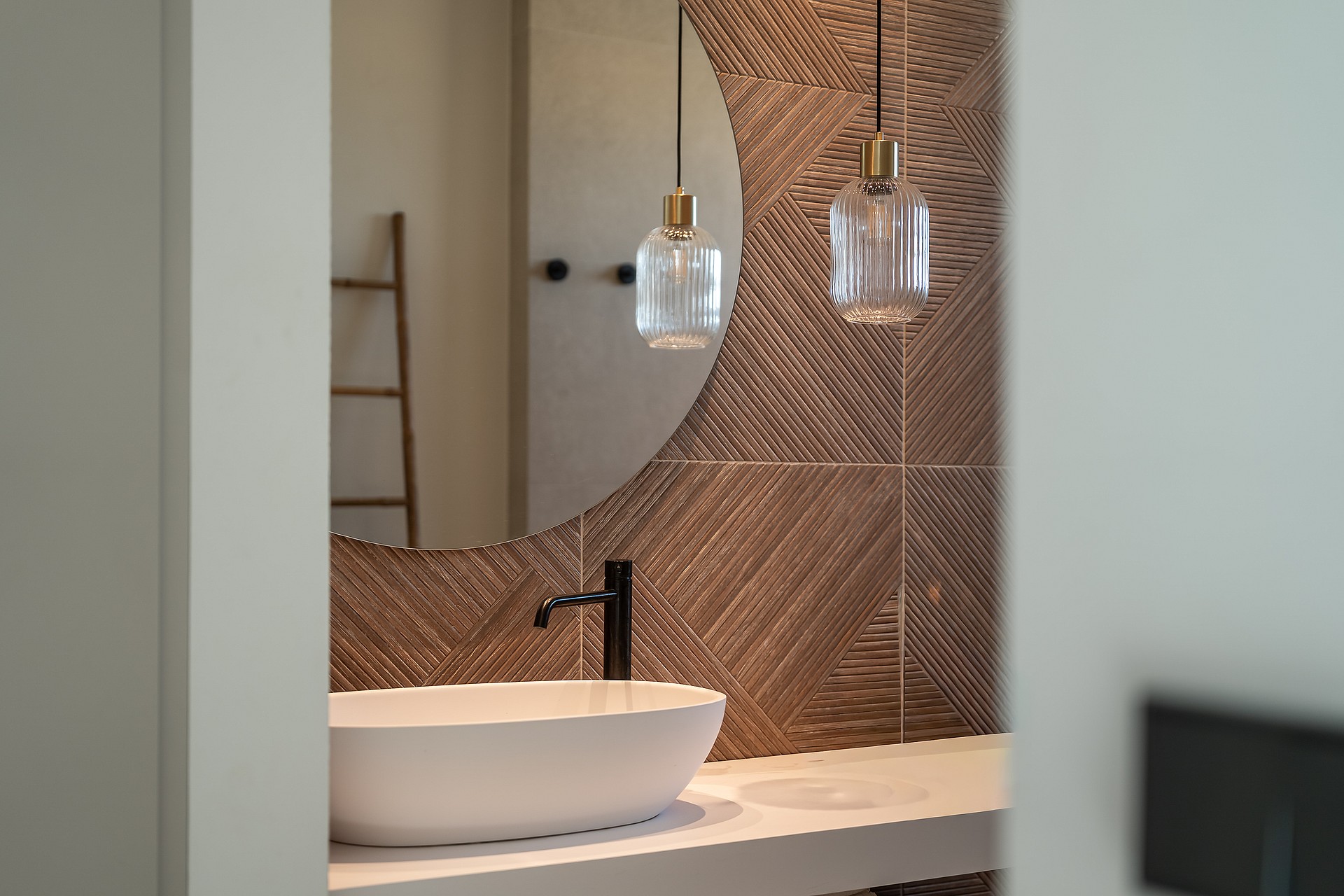 Close-up of a bathroom vanity featuring a white vessel sink, matte black faucet, and circular mirror against a textured brown tiled wall with glass pendant lights.