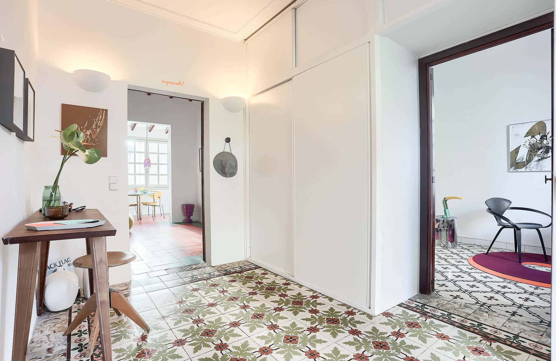 A bright hallway connecting two rooms, featuring patterned colorful tile flooring, white walls, and built-in storage closets.
