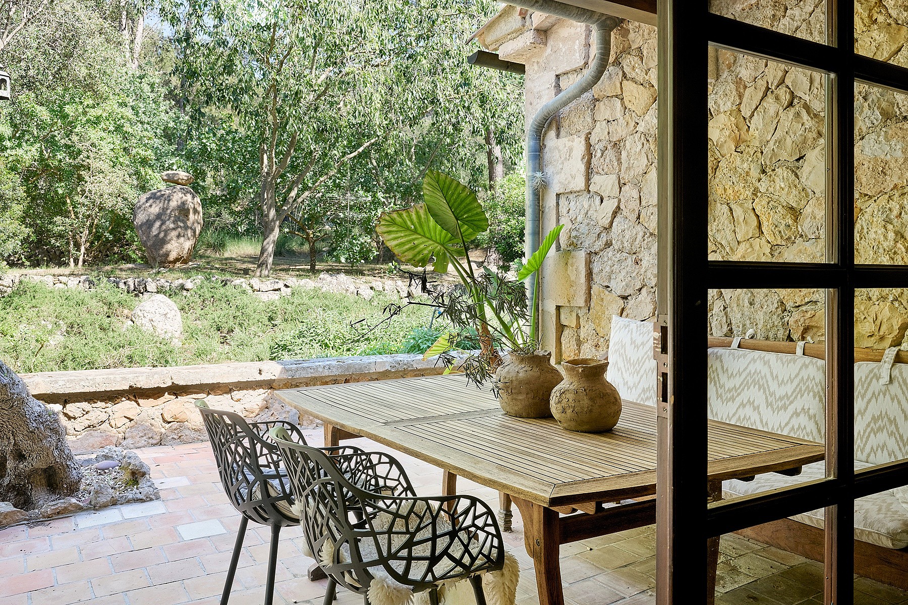 Outdoor dining set on a stone-paved terrace next to a rustic rubble stone wall, looking out to garden greenery.