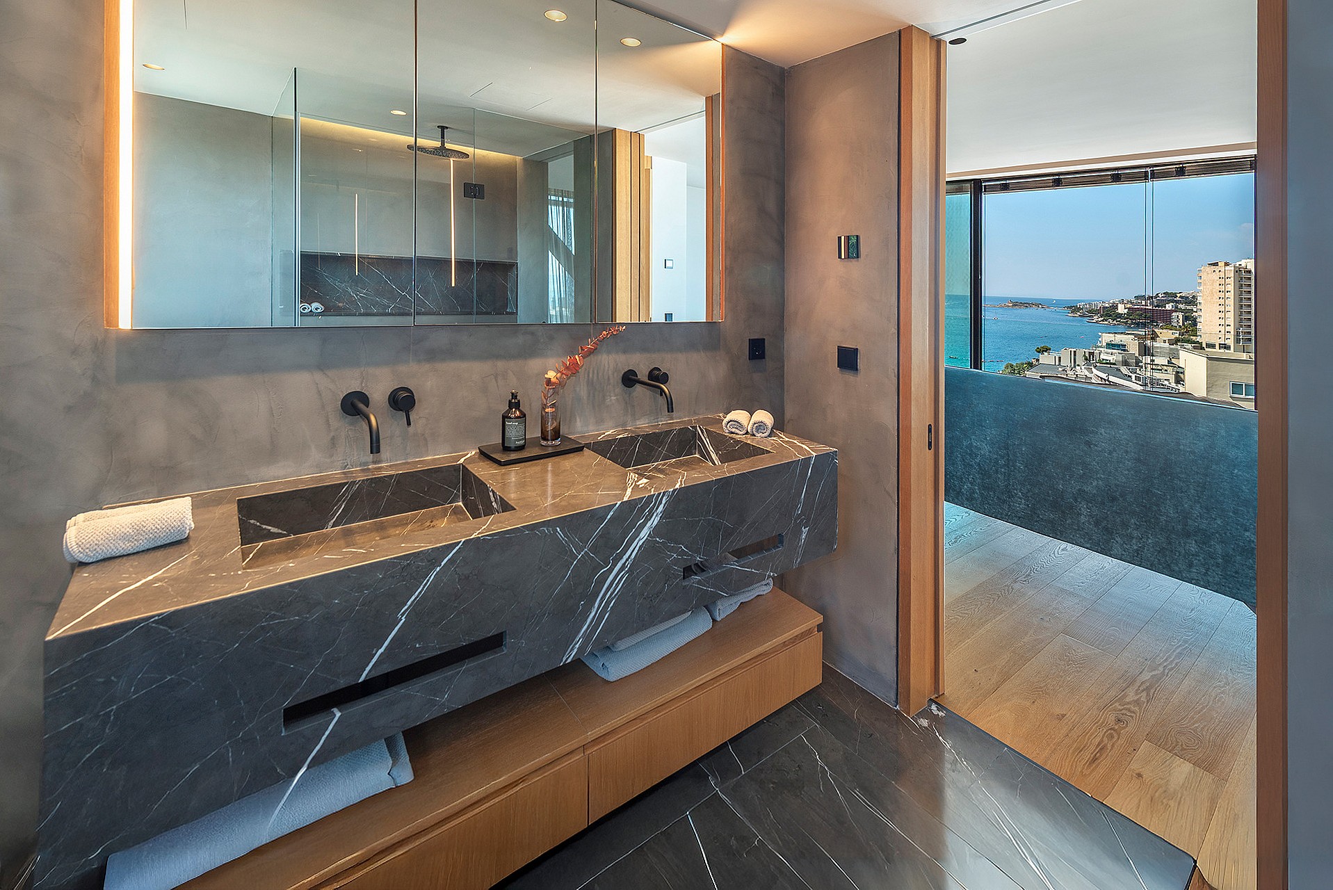 A modern bathroom featuring a grey marble double vanity with matte black faucets, a large mirror, and a view to the sea.