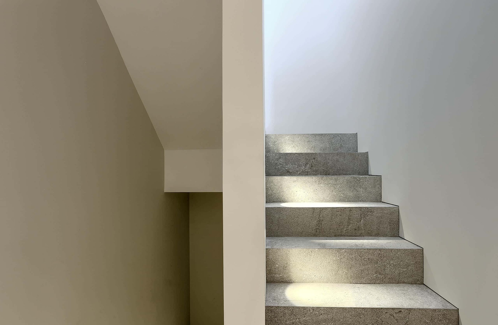 Interior staircase made of speckled grey stone with plain white walls and bright natural light from a skylight above.