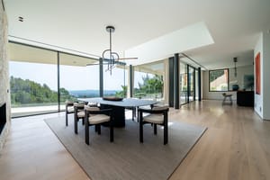 Dining room with an oval table and six chairs on a textured rug, floor-to-ceiling glass walls, and oak wood flooring.