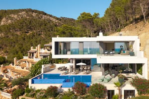 Aerial view of a white, multi-level modern villa built into a hillside, featuring a blue tiled infinity pool and sun loungers.