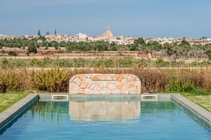 A rectangular swimming pool featuring a natural stone waterfall wall with the village of Santanyí in the background.