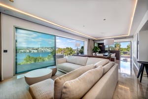 A contemporary open-plan living room featuring a large beige sectional sofa, polished stone floors, and floor-to-ceiling glass sliding doors opening to a lush garden view.