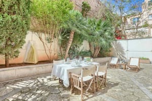 An outdoor dining table with white tablecloth and director chairs set on a gravel terrace shaded by mature trees.