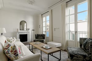 Bright living room with cream furnishings, wooden fireplace mantle, tall windows overlooking the city, and herringbone wooden floors