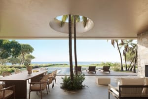 Outdoor dining and lounge area on a shaded terrace featuring a circular opening in the ceiling with a tree, overlooking a pool and the sea.