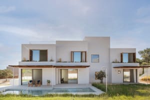 Modern white two-story villa with wooden shutters and a rectangular pool reflecting the sky, bordered by green grass.