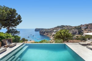 An infinity edge swimming pool with turquoise mosaic tiles overlooking a sea bay with rocky cliffs and pine trees.