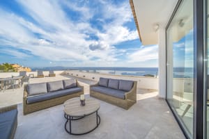 A wide outdoor terrace featuring woven lounge furniture, stone flooring, and expansive views of the sea under a blue sky.