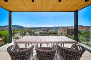 A covered terrace with a wood-paneled ceiling, a long dining table, and dark woven chairs overlooking a landscape of mountains.