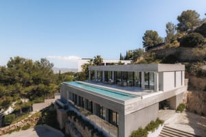 Elevated view of a contemporary concrete villa featuring a rooftop infinity pool and floor-to-ceiling glass walls.