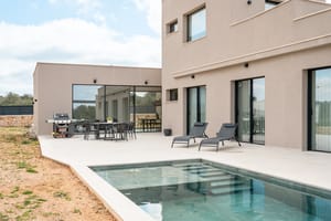 Rear facade of a modern beige villa showing a stone terrace, swimming pool with steps, and outdoor dining area.
