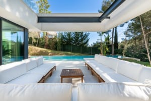 White cushioned outdoor seating under a steel pergola roof beside the swimming pool and pine garden