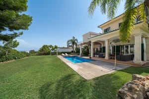 Exterior view of a villa with a rectangular swimming pool, manicured lawn, sandstone columns, and a shaded terrace.