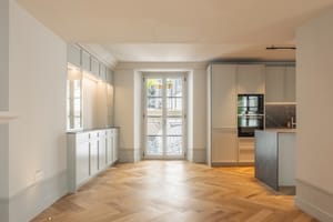 Interior view of a renovated apartment featuring light oak herringbone flooring, contemporary kitchen cabinetry, and French doors.