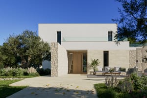 Modern villa exterior with white render and natural stone cladding, featuring a terrace area and olive trees under bright sun.
