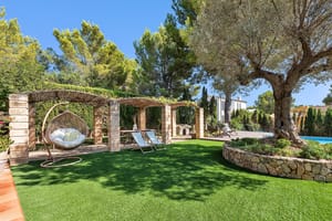 A green manicured lawn with a stone pergola, hanging wicker swing chair, and an mature olive tree in a sunny garden.