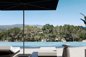 A poolside terrace with white sun loungers, a black parasol, and a clear view of pine trees and distant mountains.