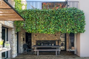 A shaded outdoor dining terrace with stone walls, a wooden table, bench seating, and a large climbing ivy wall.
