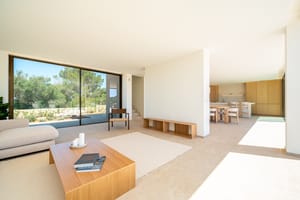 A bright, open-plan living room with a light beige sofa, wooden coffee table, and large sliding glass doors overlooking trees.