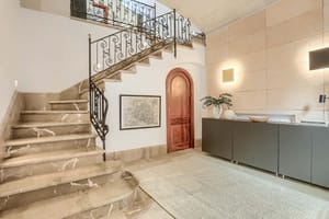 An interior shot of an entrance hall featuring a wide marble staircase with a wrought-iron railing, a curved wooden door, and a textured stone feature wall.