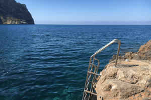 A stainless steel swim ladder attached to natural rocky coastline leading into deep blue Mediterranean sea waters.