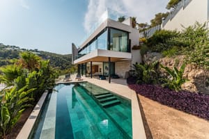 Modern two-story villa with glass walls and stone terracing, set against a rocky hill with an infinity-edge swimming pool.