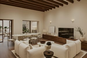 Open living and dining room with pale sofas, oval travertine table, walnut media unit and exposed wooden beams, glazed doors to a terrace and pool.