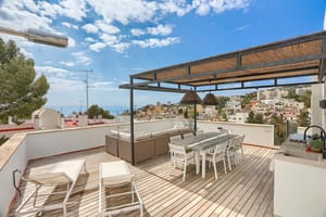 A spacious outdoor rooftop terrace featuring a wooden deck, a covered dining area with a woven pergola, lounge seating, and distant sea views over a coastal neighborhood.