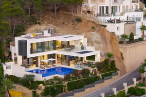 Aerial view of a white modern villa carved into a cliffside, featuring a swimming pool, multiple terraces, and glass facade.