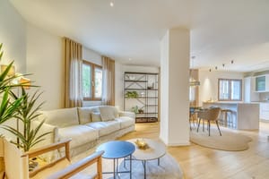 Bright open-plan living room with a beige sofa, round coffee tables, wooden flooring, and adjacent dining and kitchen areas.