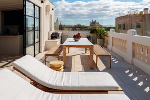 An expansive outdoor rooftop terrace featuring white sun loungers, a wooden dining table, and a view over the Santa Catalina skyline under a clear blue sky.