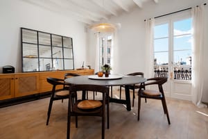 Round dark wood dining table with woven-seat chairs beneath a white beamed ceiling, French doors opening to rooftop views.