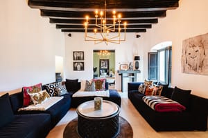 Interior view of a living room with dark low-profile sofas, a central round metal coffee table, and an ornate brass chandelier hanging from dark exposed ceiling beams.